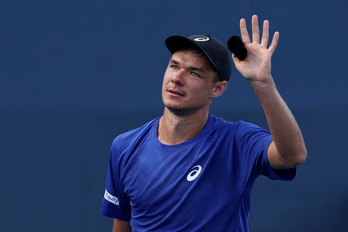 Kamil Majchrzak in sports attire waves, wearing a cap and branded t-shirt, likely a tennis player acknowledging the crowd