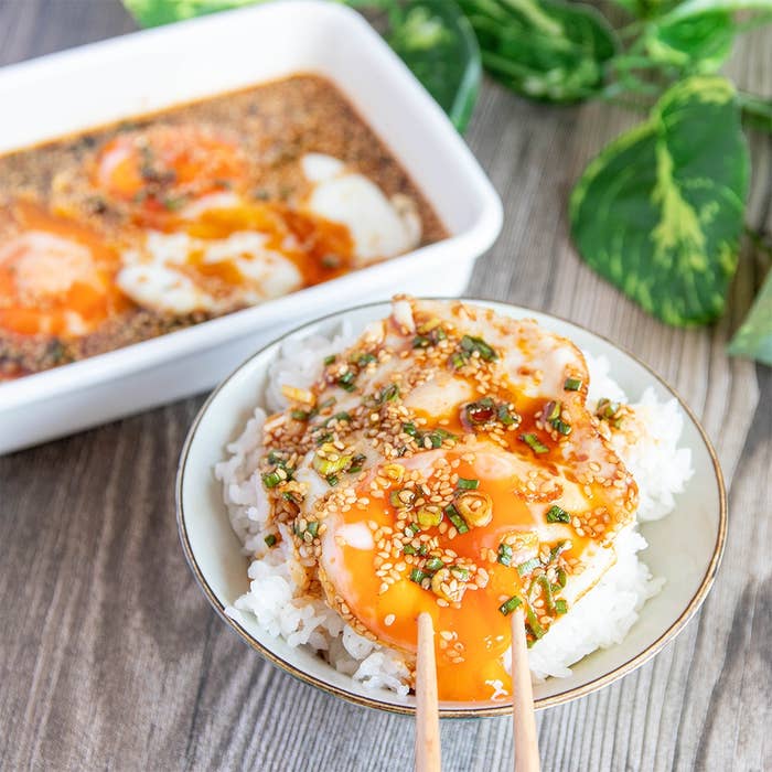 温かいご飯の上に半熟卵が乗せられ、ねぎとごまが散らされている料理。背景にオーブン皿と観葉植物がある。