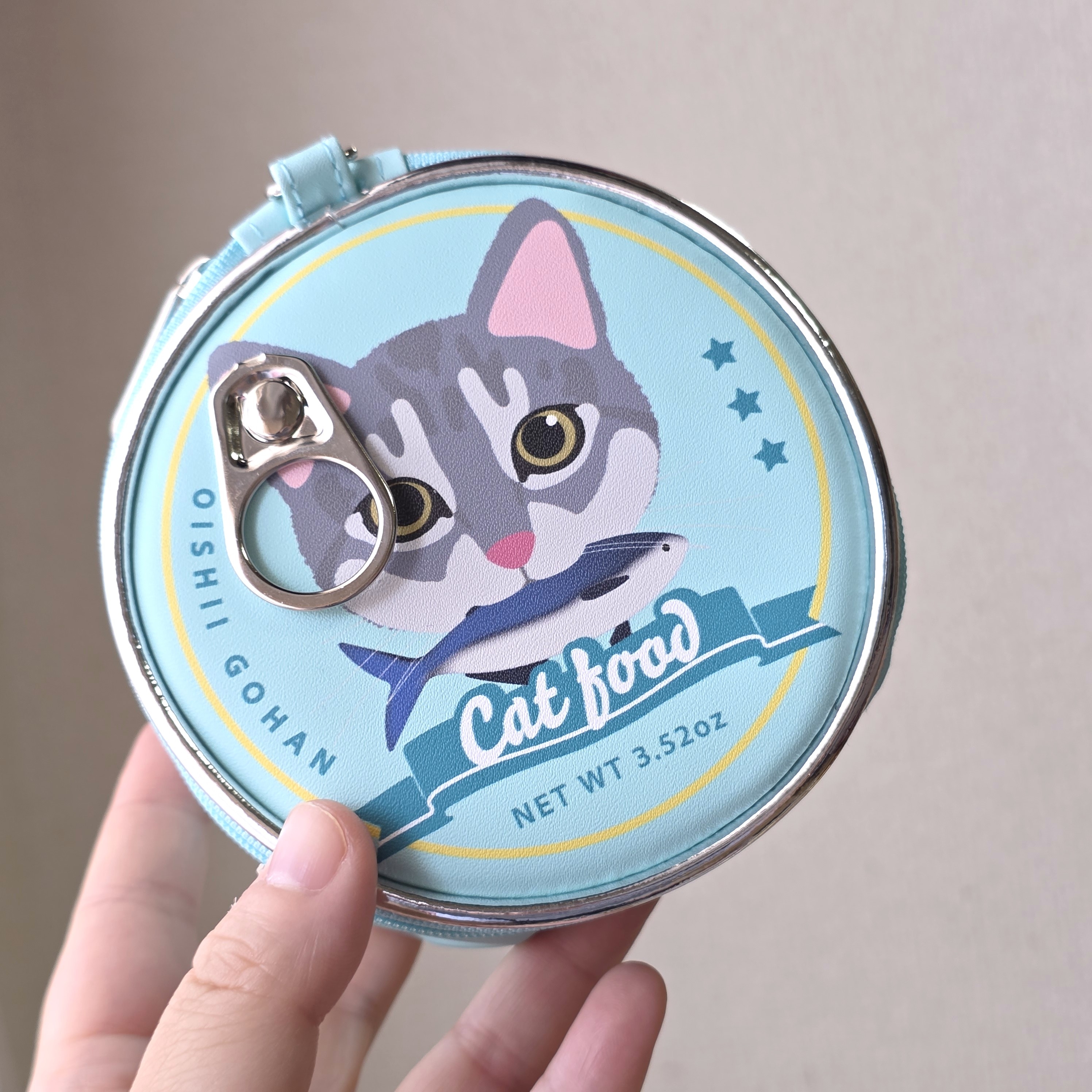 猫のイラストが描かれた丸い猫缶デザインのコインポーチ。手で持たれている。