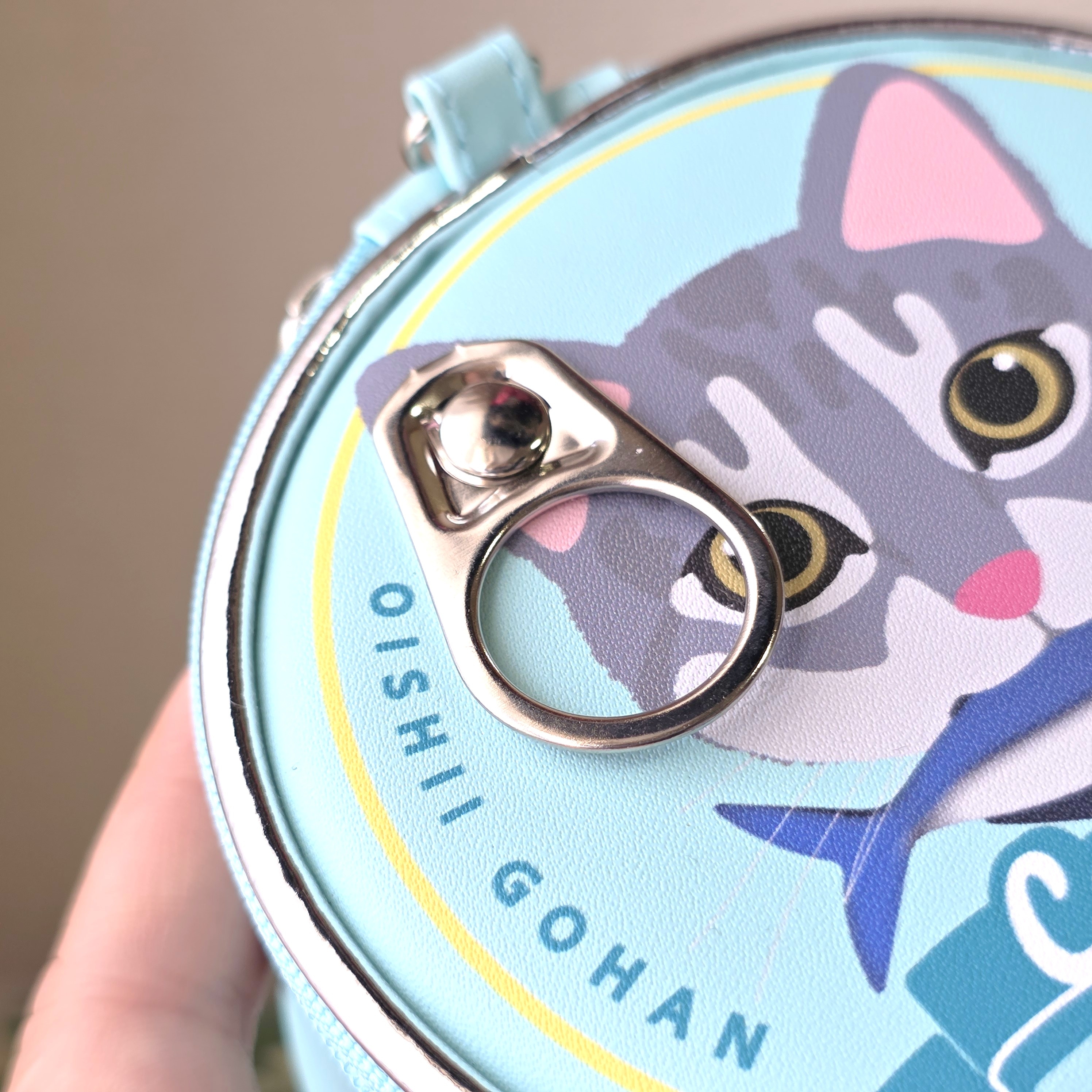 猫のイラストが描かれた円形の缶。テキストは「OISHII GOHAN」。プルタブが見える。