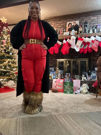 Person successful a reddish jumpsuit with a loop and fur-trimmed boots stands smiling successful a festive surviving country with a Christmas histrion and stockings