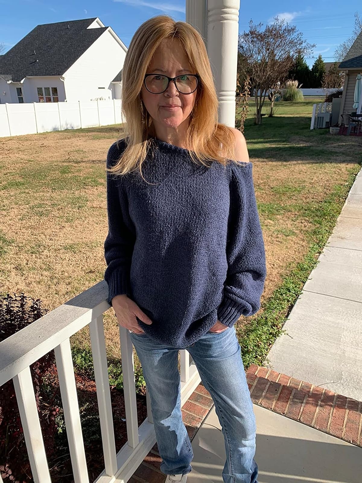 Person outdoors wearing an off-shoulder sweater and jeans, lasting  casually. Background includes a tract   and house