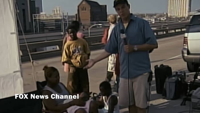 News newsman successful a bluish garment stands connected a thoroughfare with a microphone, gesturing to adjacent seated people