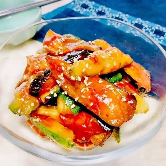 透明なボウルに盛られた甘辛ソースの茄子料理。ゴマが振りかけられ、色鮮やかな見た目。