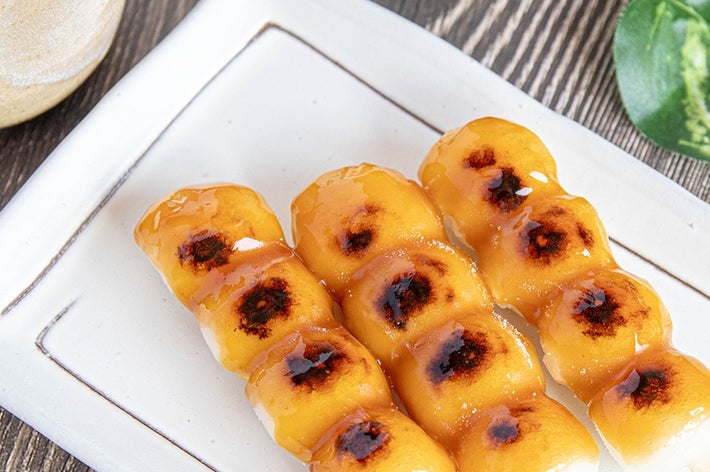 三本の焼き団子が皿に並ぶ。団子は焦げ目がついており、そばには飲み物。緑の葉が背景に広がっている。