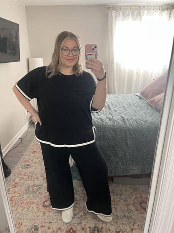 Person taking a mirror selfie in a relaxed, casual matching black top and pants with white trim, standing in a cozy bedroom