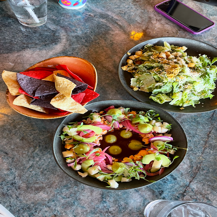 Three dishes on a table: chips, a salad with croutons, and a ceviche garnished with pickled onions and herbs. A drink and a phone are nearby