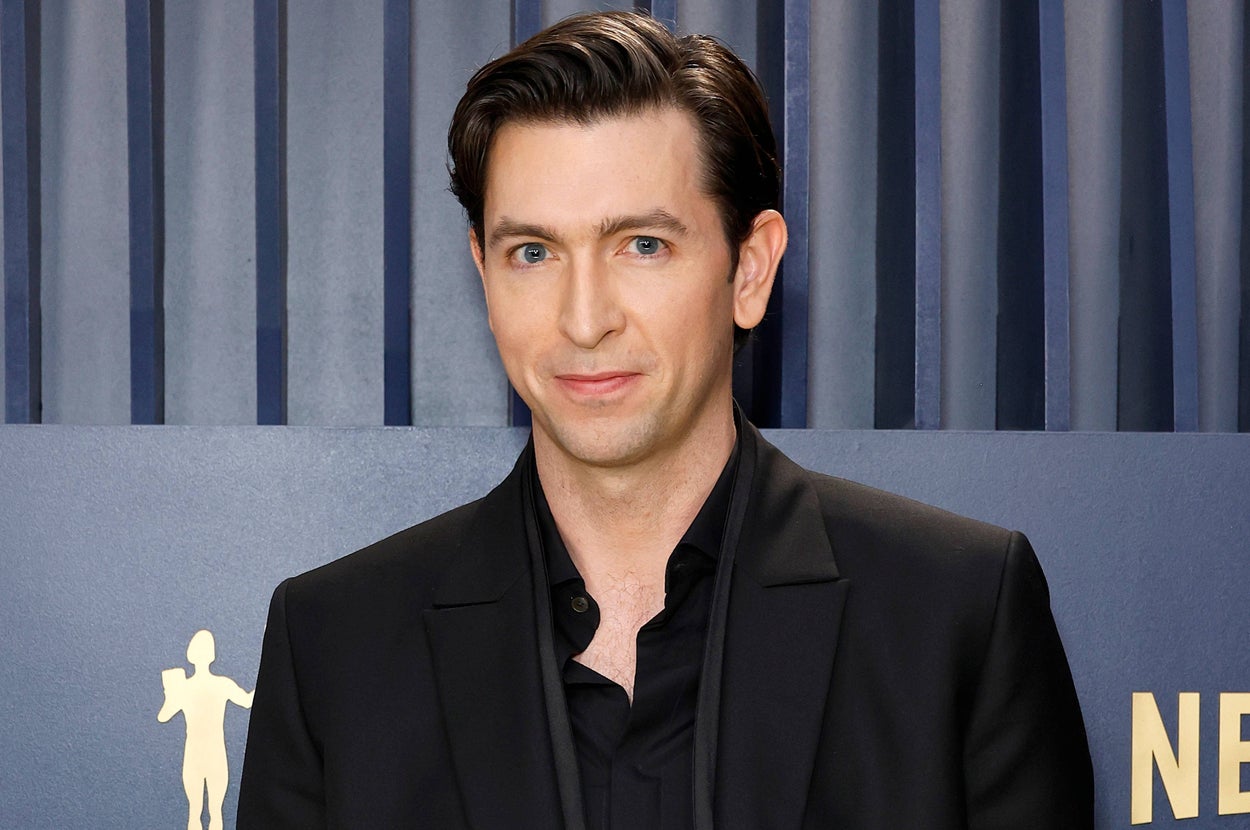 A person on the red carpet in a sleek black suit with a dark shirt, standing against a backdrop with award logos