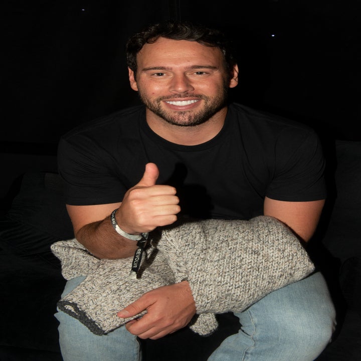 Smiling man seated, wearing a black T-shirt and holding a gray knit sweater, gives a thumbs-up gesture
