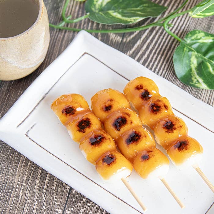 陶器の皿に盛られたみたらし団子の串3本。葉の飾りと湯呑が背景に置かれている。