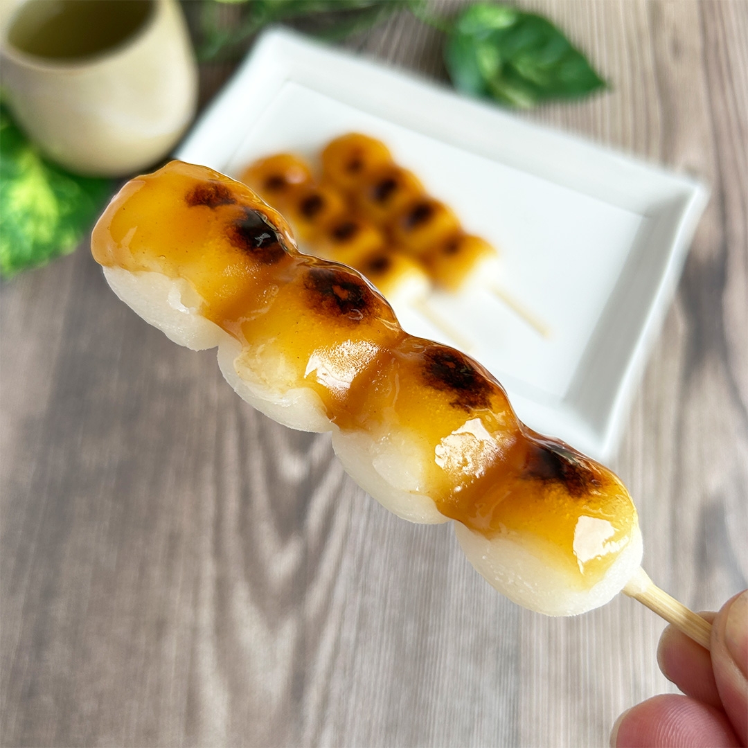 手で持たれたみたらし団子の串。背景に白い皿と他の団子。