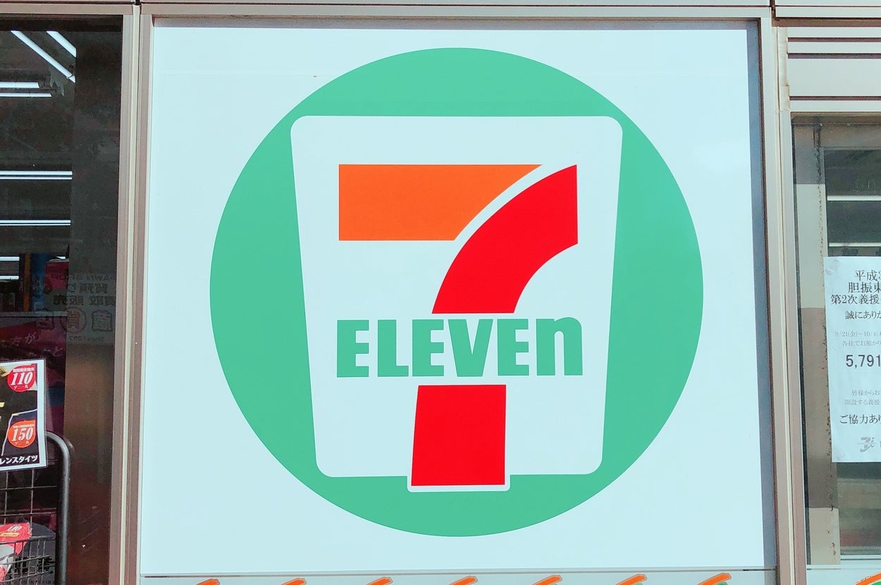 セブンイレブンの大きなロゴが店舗のガラス窓に描かれている。周囲に商品陳列が見える。
