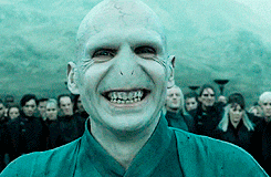 A character with pale skin and no hair grins menacingly, surrounded by people standing in the background