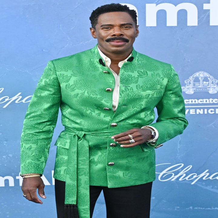 Person in a patterned blazer with a sash and dark pants on a blue carpet at a film event backdrop
