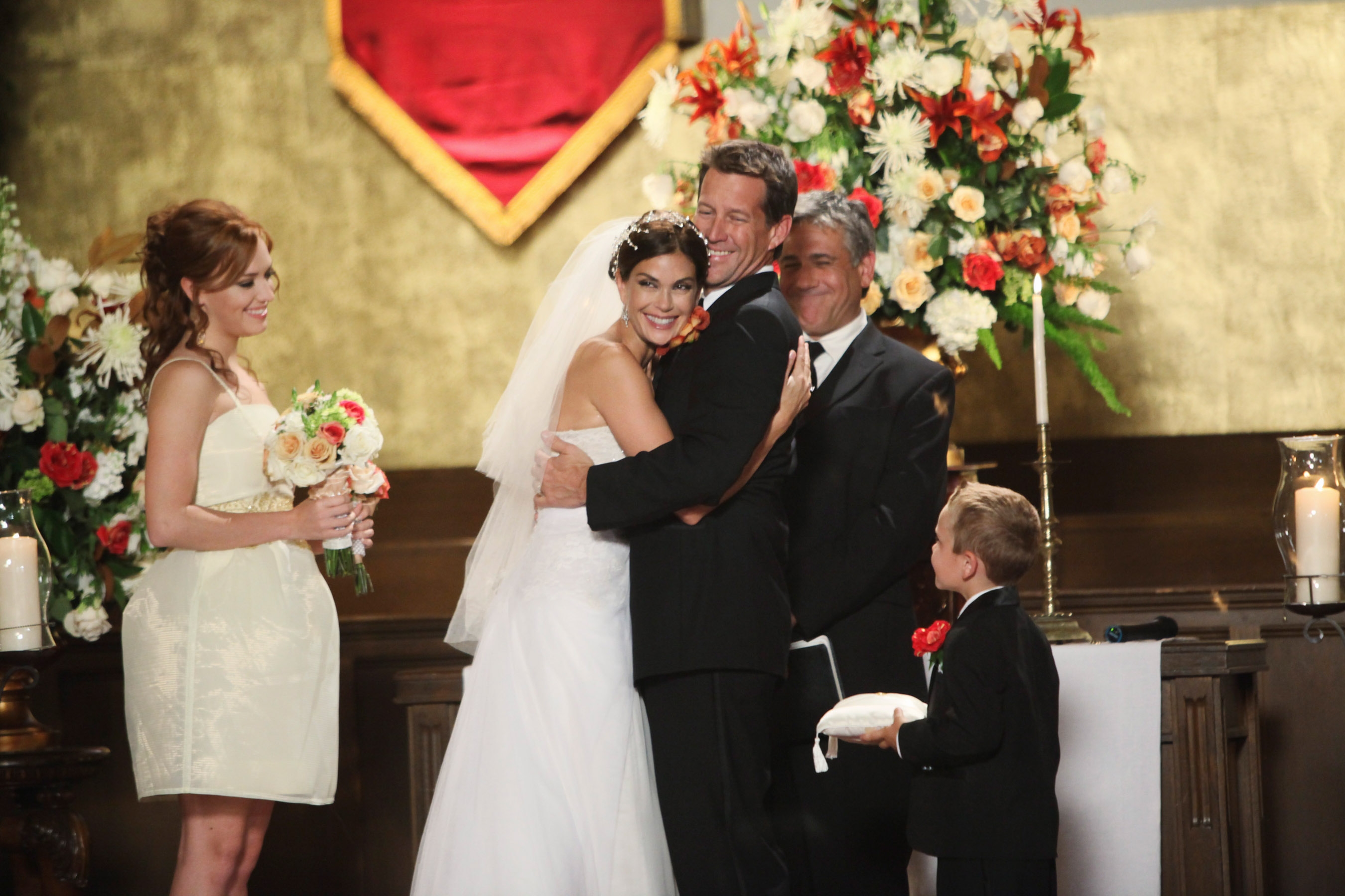 Bride in a strapless gown hugs groom beside bridal party in a church setting. The scene is joyful, with floral decorations in the background