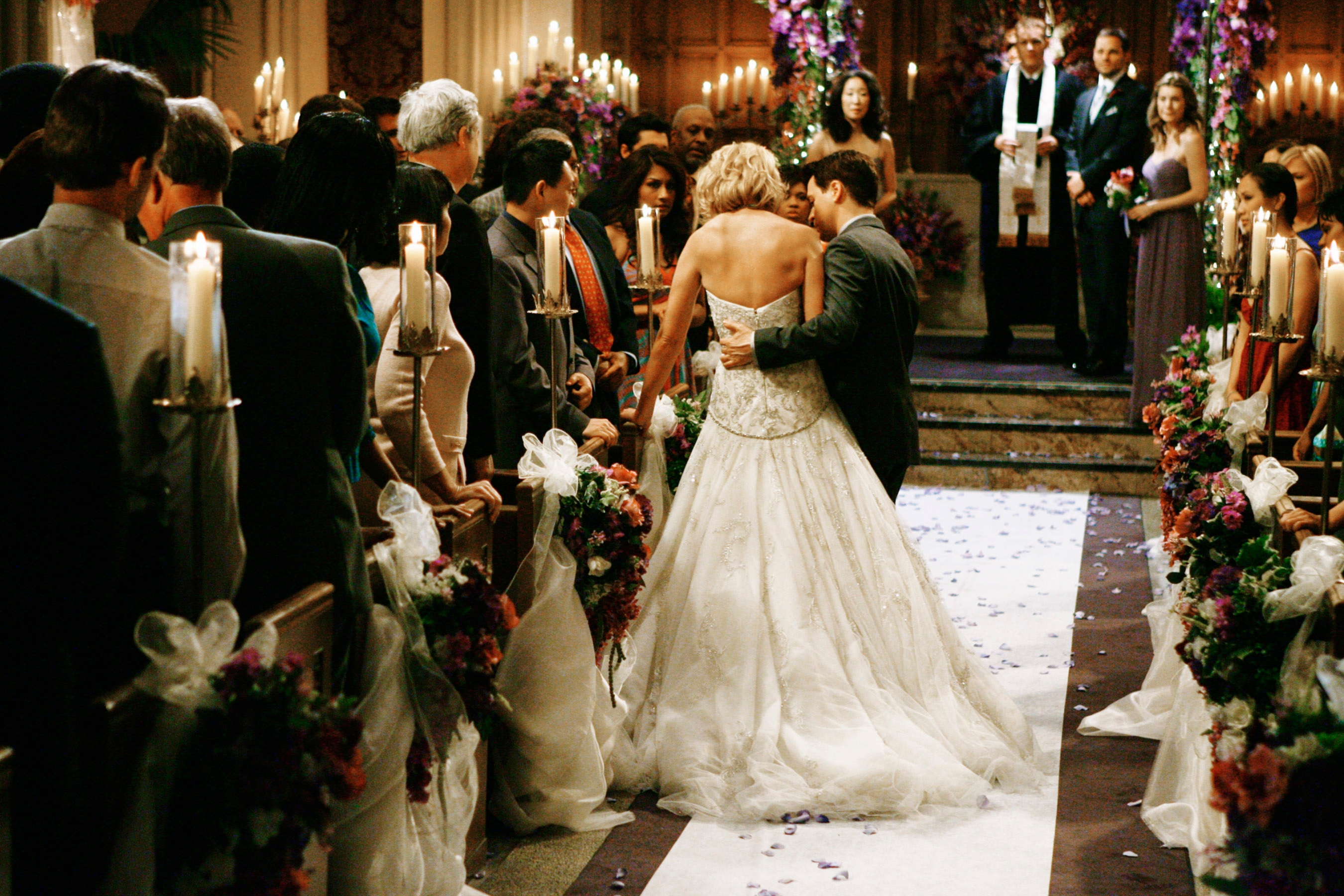 Bride and groom walk down the aisle in an ornate church setting, surrounded by guests. The bride wears a strapless gown; the groom is in a classic suit