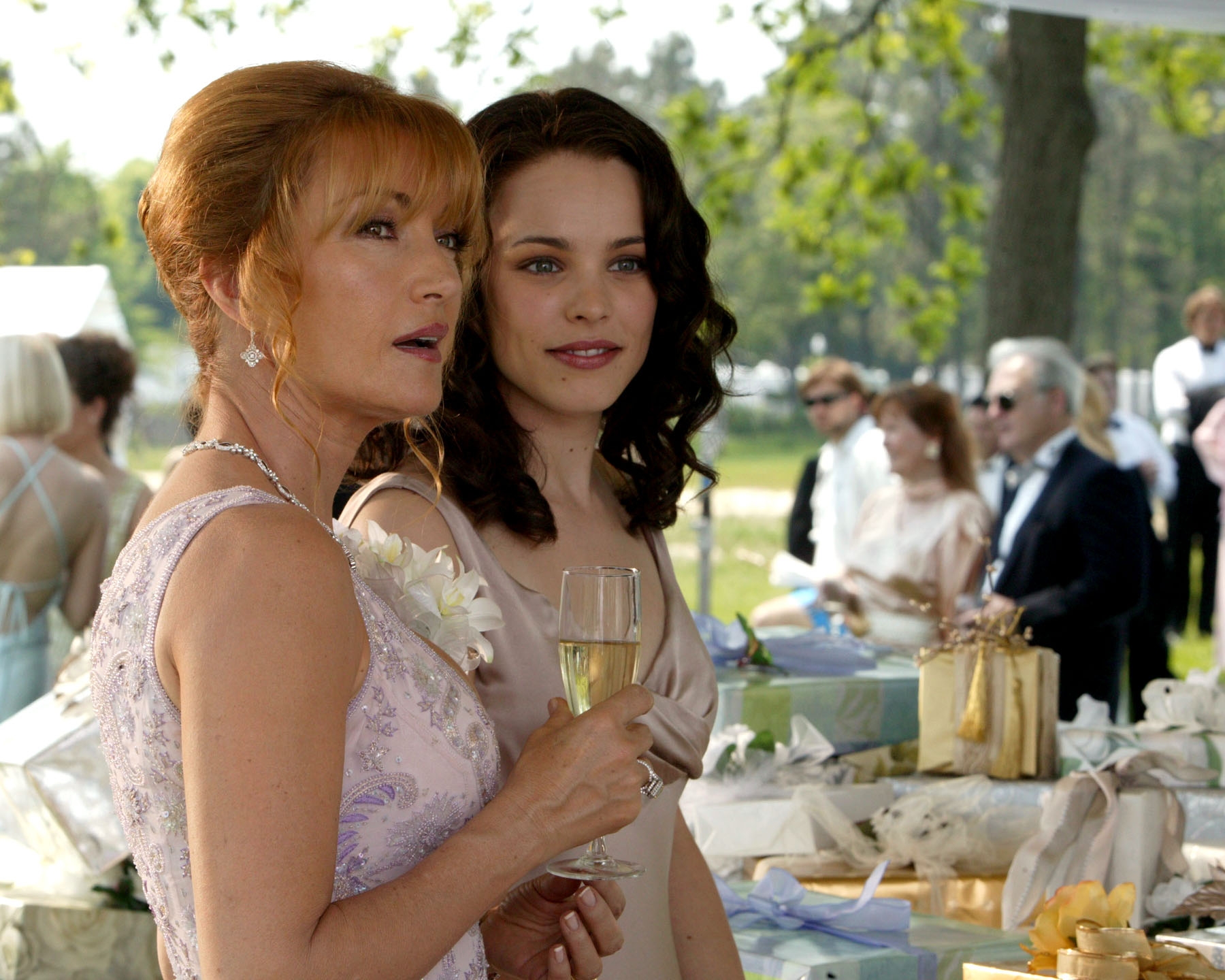 Two women in elegant dresses at a wedding reception, holding champagne flutes, surrounded by gift tables and guests in the background