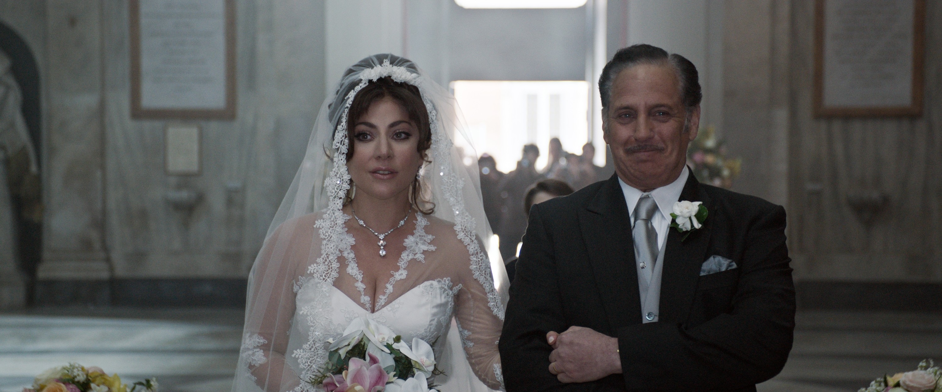 Bride in lace gown with floral veil stands arm-in-arm with older man in suit, smiling, at a wedding ceremony