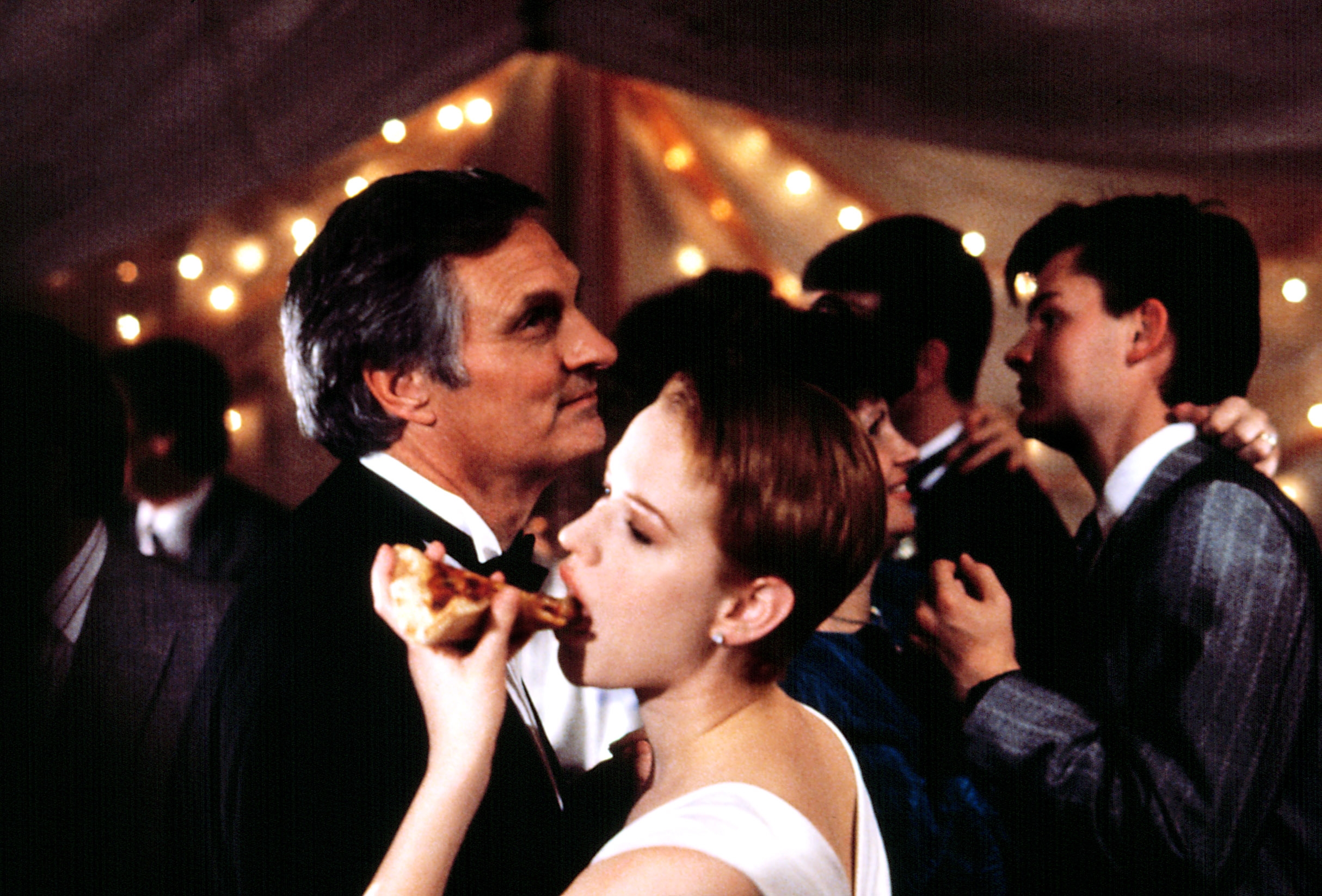 A person in formal attire eats pizza at a wedding party, surrounded by guests in suits and dresses under string lights