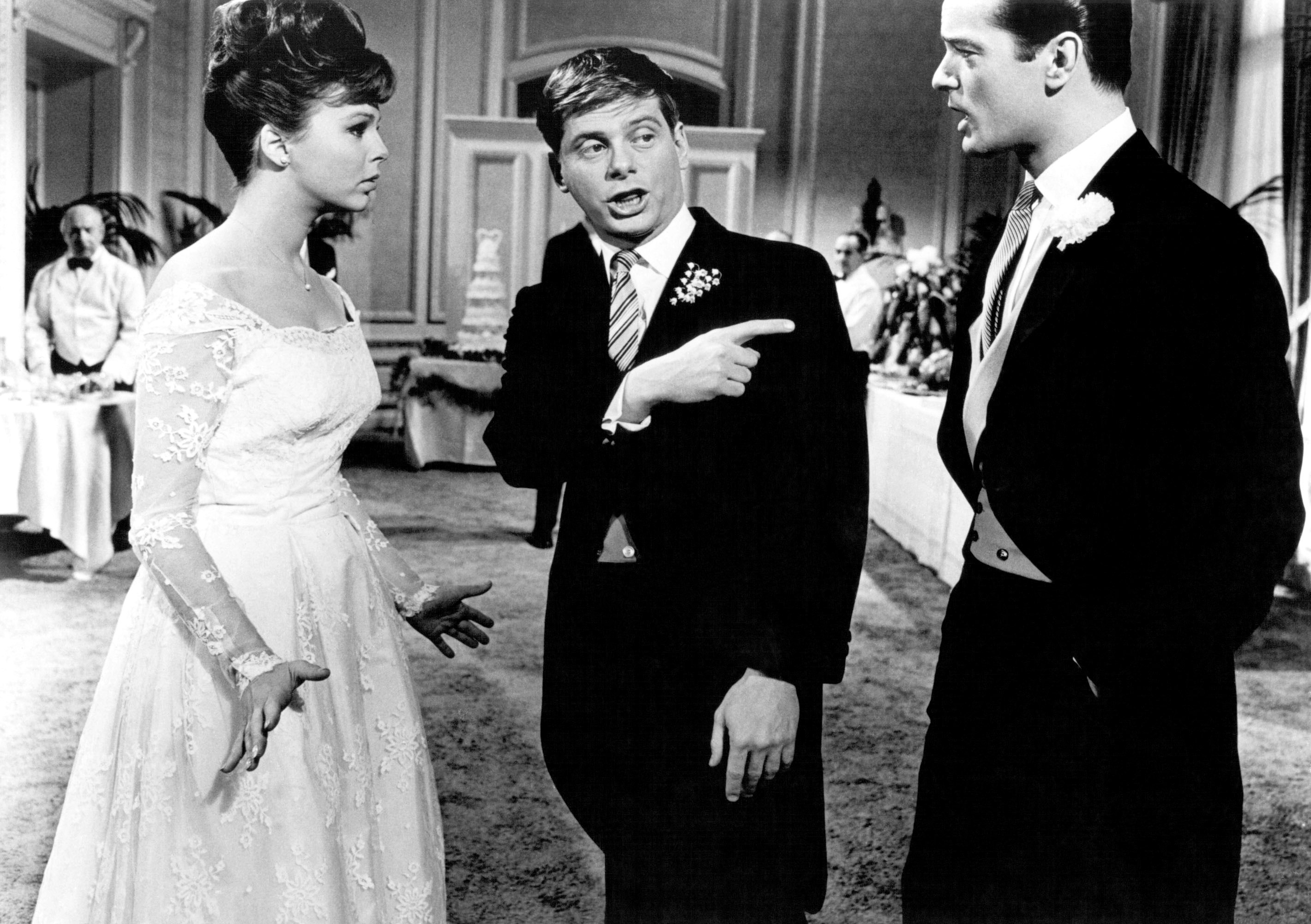 A bride in a classic wedding gown discusses animatedly with two men in formal suits at a wedding reception