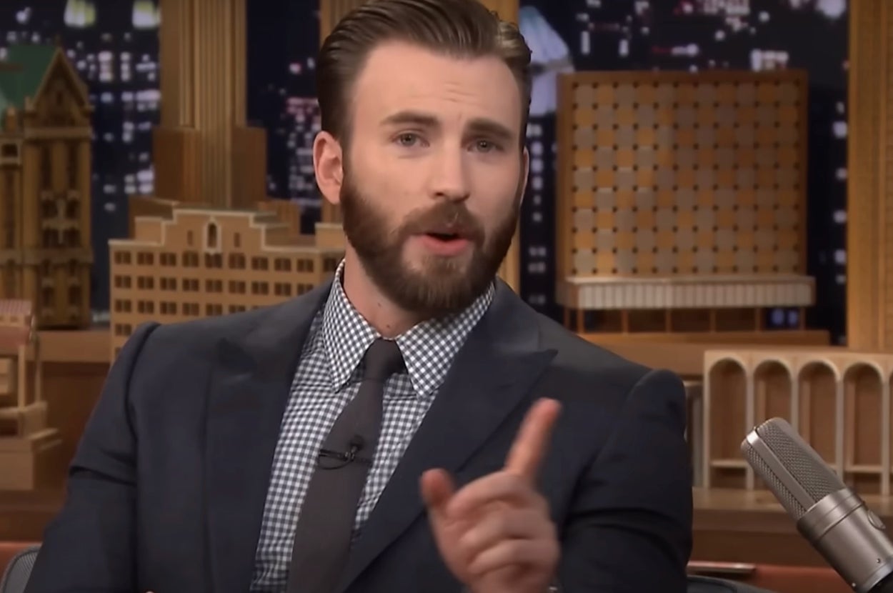 A person in a checkered shirt and dark suit gestures while speaking during a talk show interview
