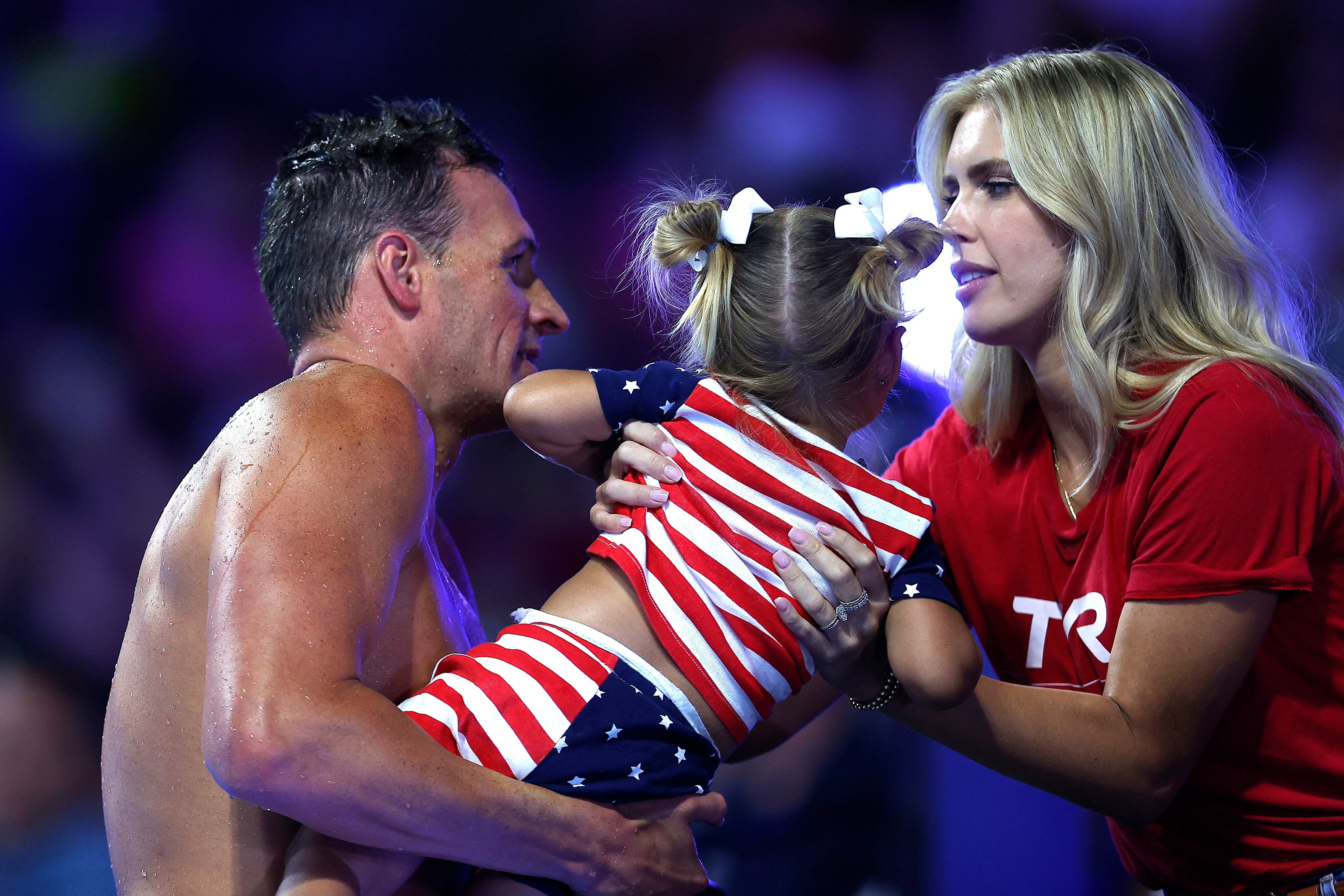 Man holds a girl wearing a star-patterned dress, while a woman adjusts her. They appear to be at a sports event
