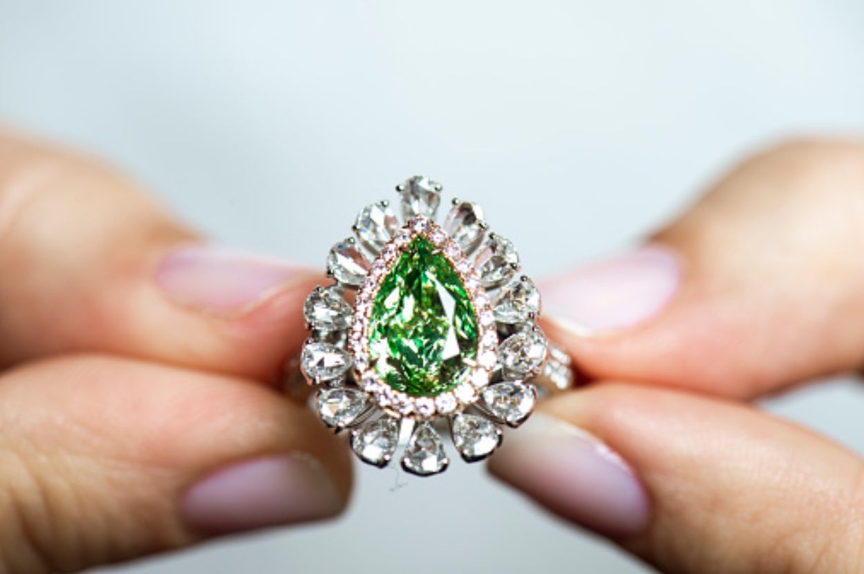 Hands holding an ornate ring with a large, pear-shaped green gemstone surrounded by clear stones