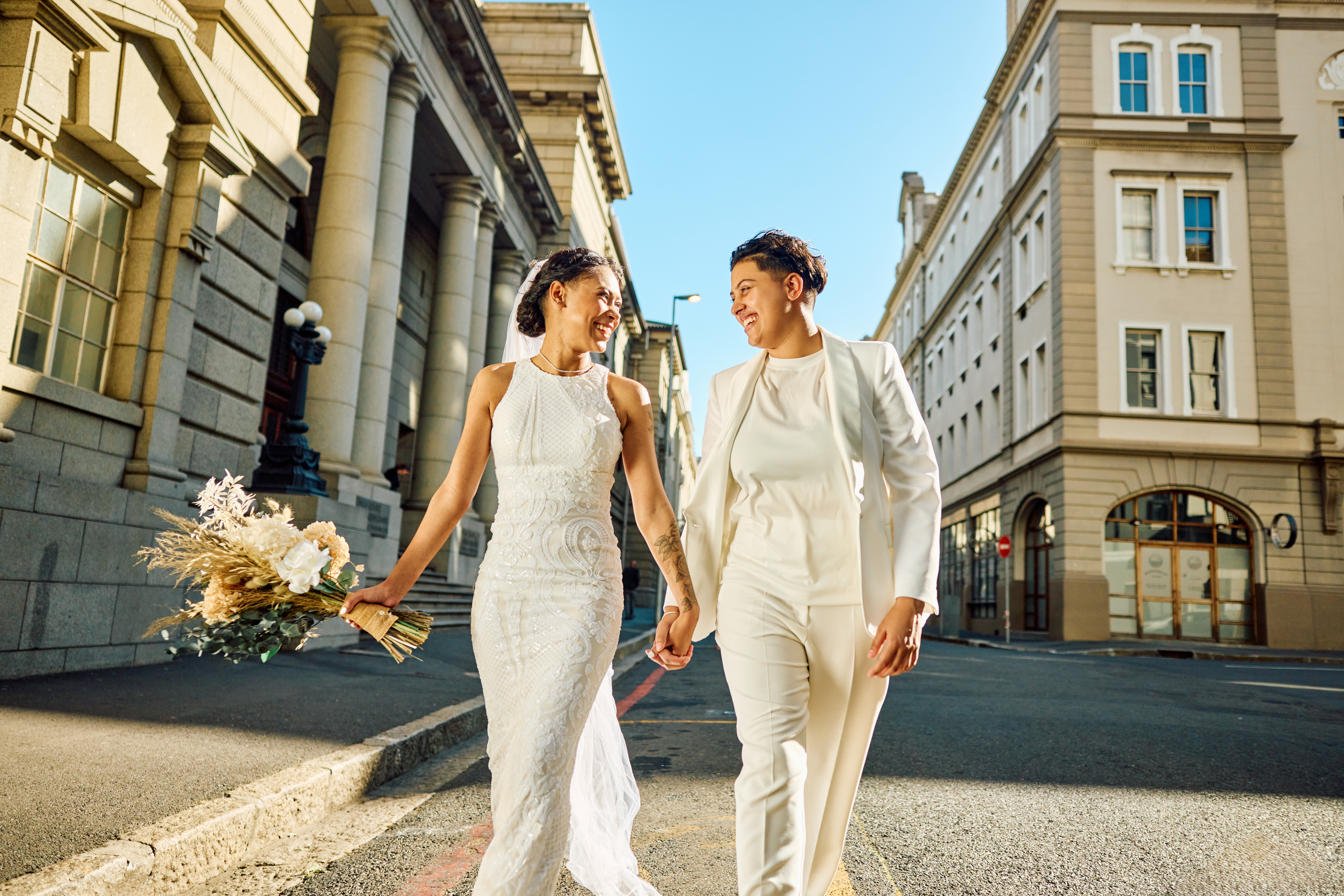 To personer som smiler og holder hender på en bygate. Den ene har på seg en blonderkjole, den andre en skreddersydd hvit drakt, og holder en bukett blomster