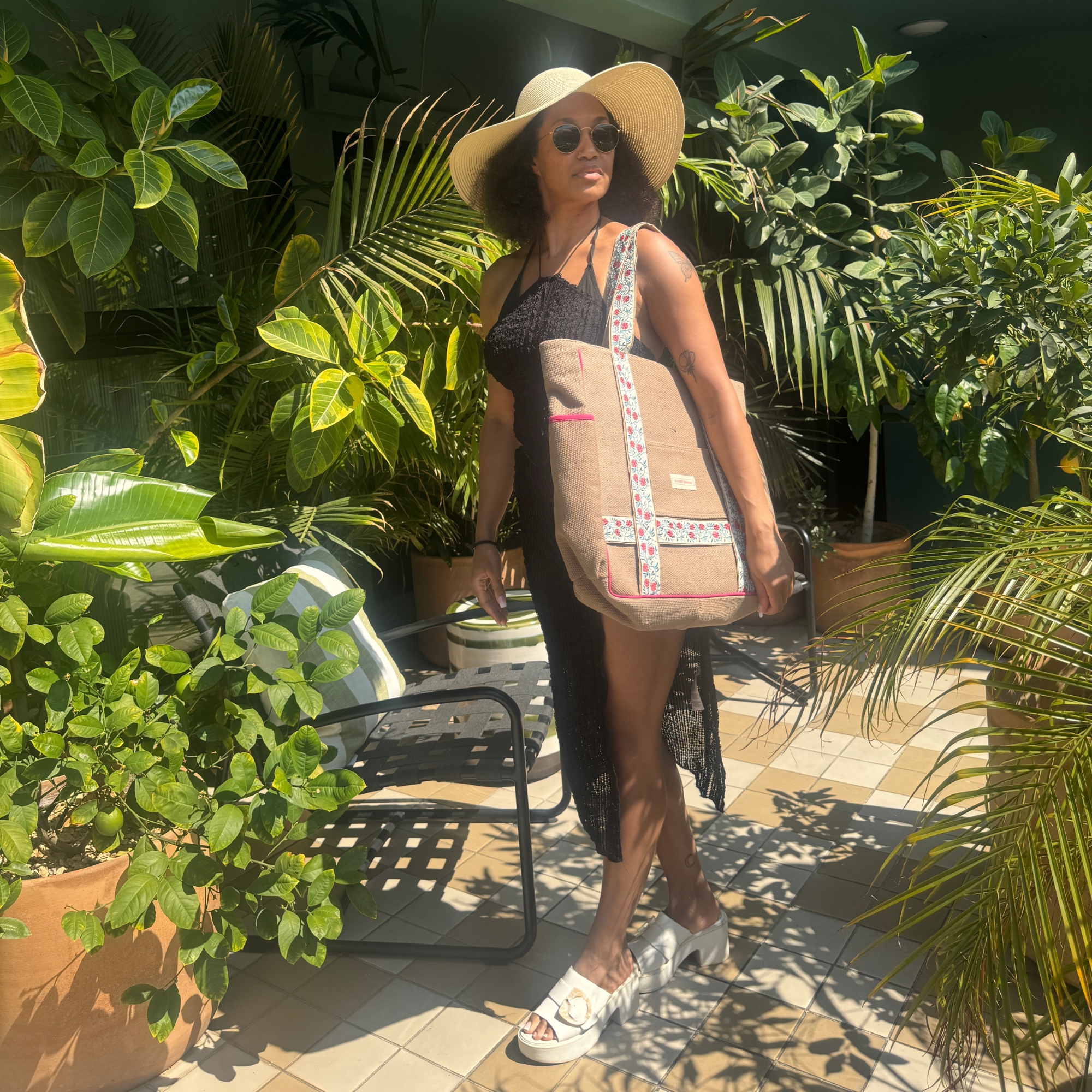 person in sun hat, sheer black dress, and white sandals stands by lush plants, holding a large patterned tote bag
