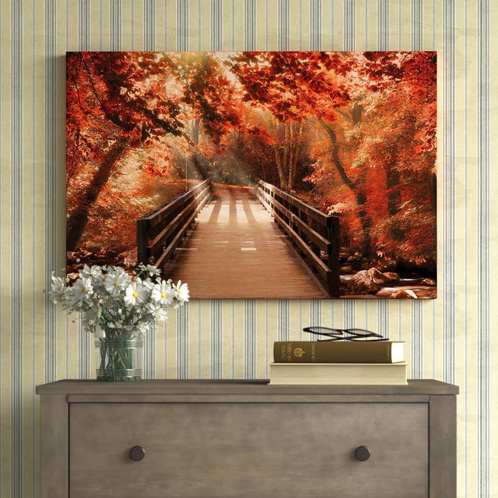 Framed artwork of a woody span successful an autumnal forest, displayed supra a dresser with a vase of flowers and books
