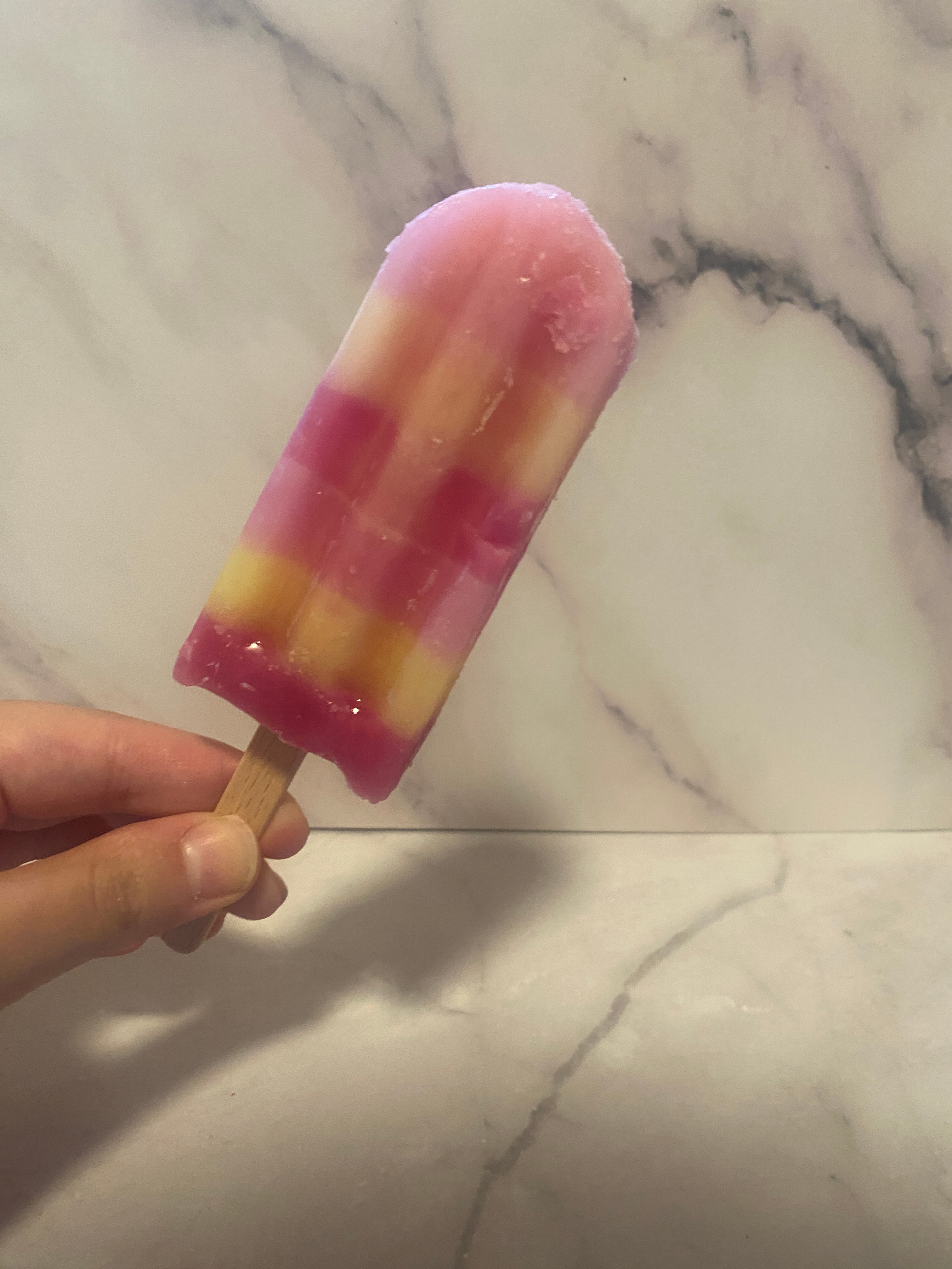 Person holding a multi-layered popsicle with pink and yellow stripes against a marble backdrop