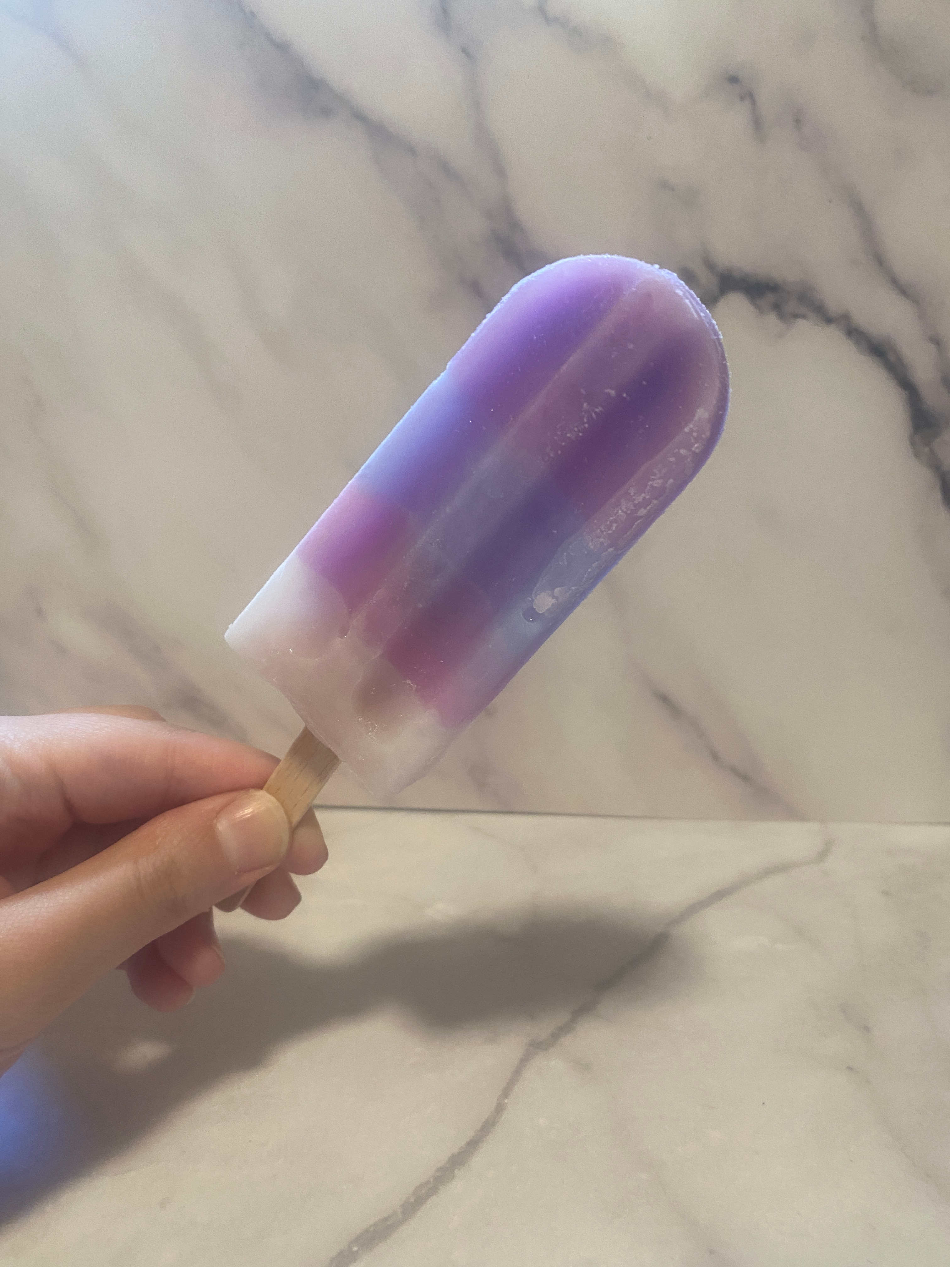 A hand holding a purple and white ombré popsicle against a marble background