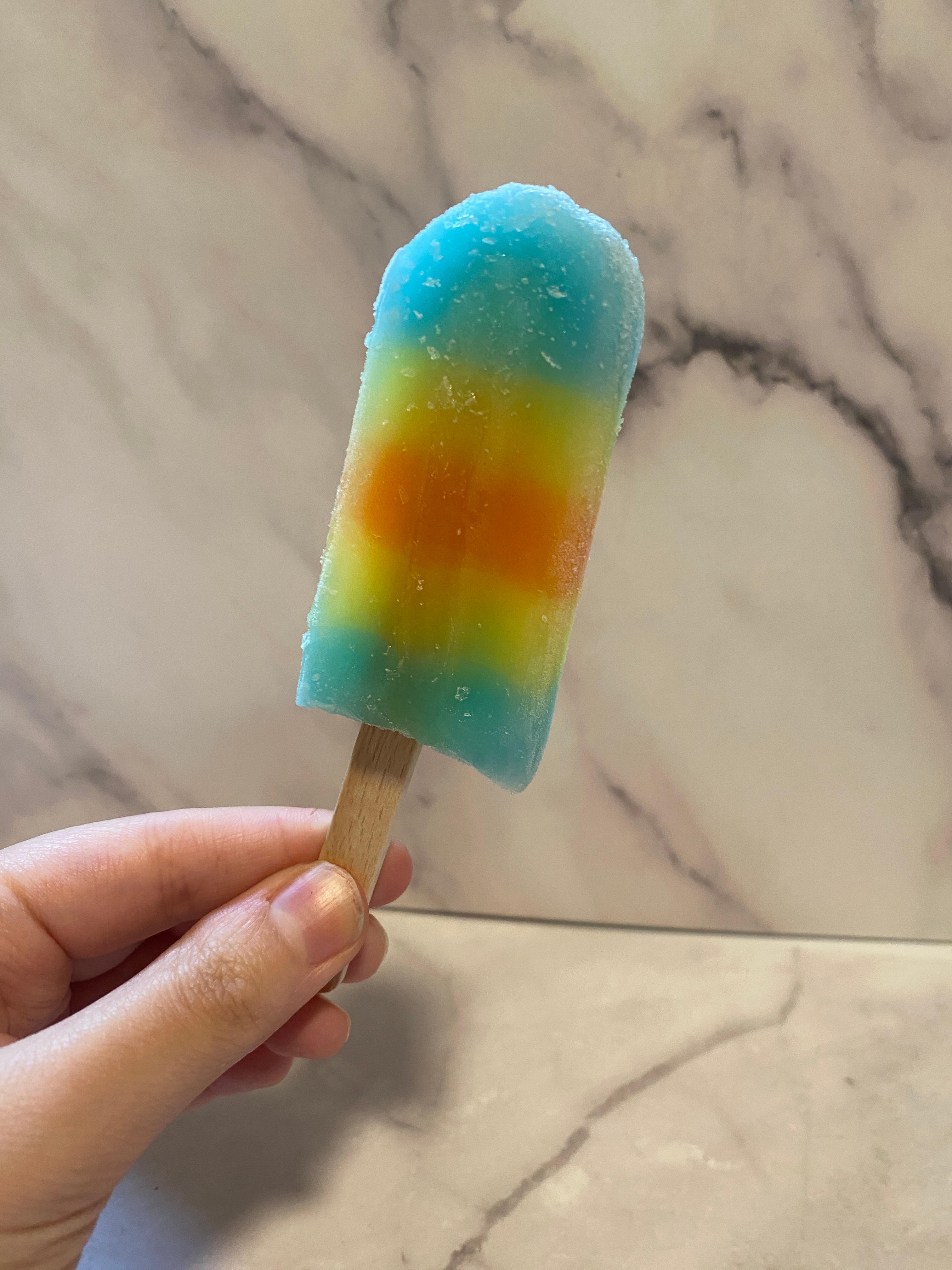 Hand holding a popsicle with blue, yellow, and orange sections against a marble background