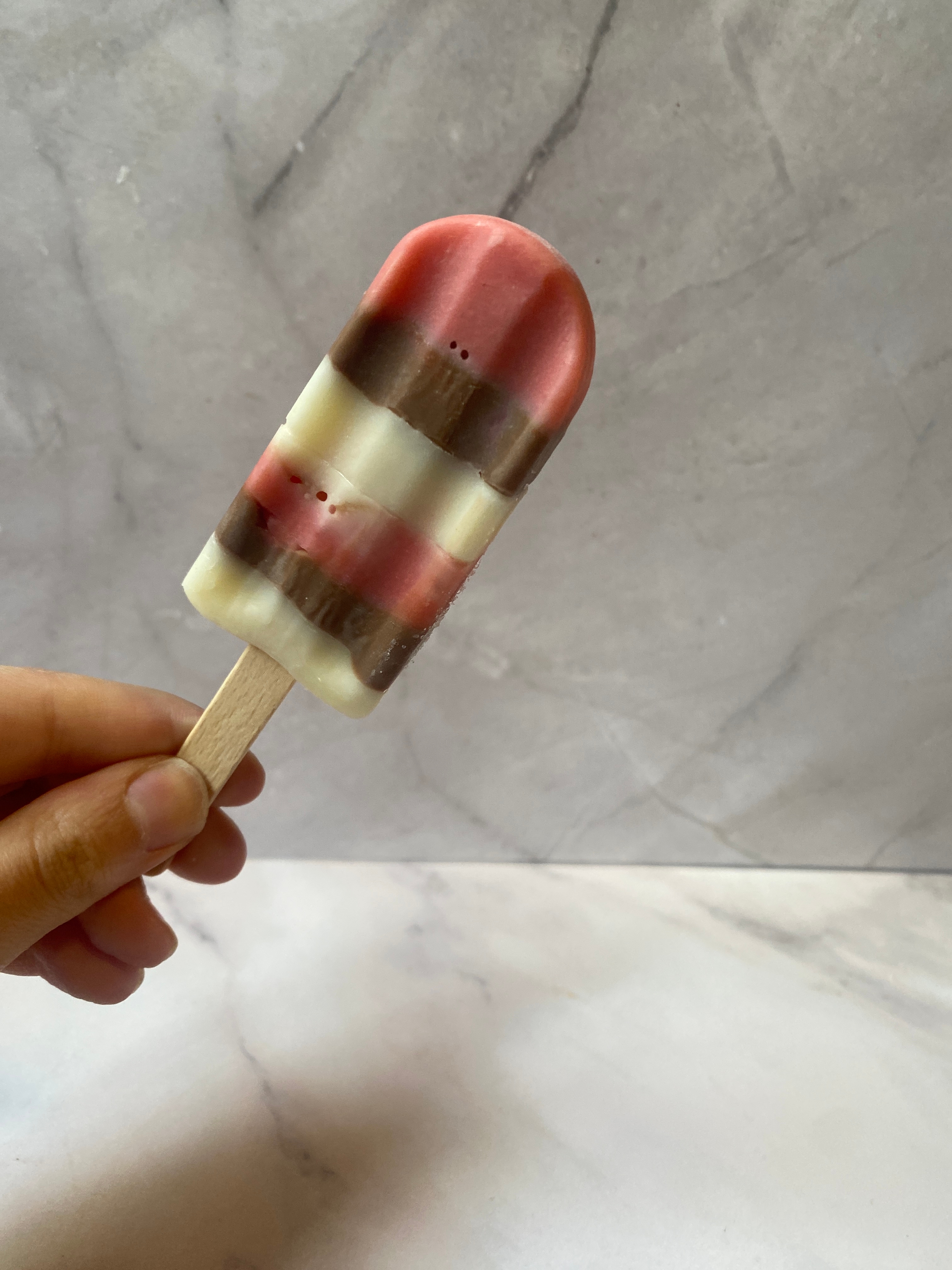 A hand holds a layered popsicle with pink, white, and brown stripes against a neutral background