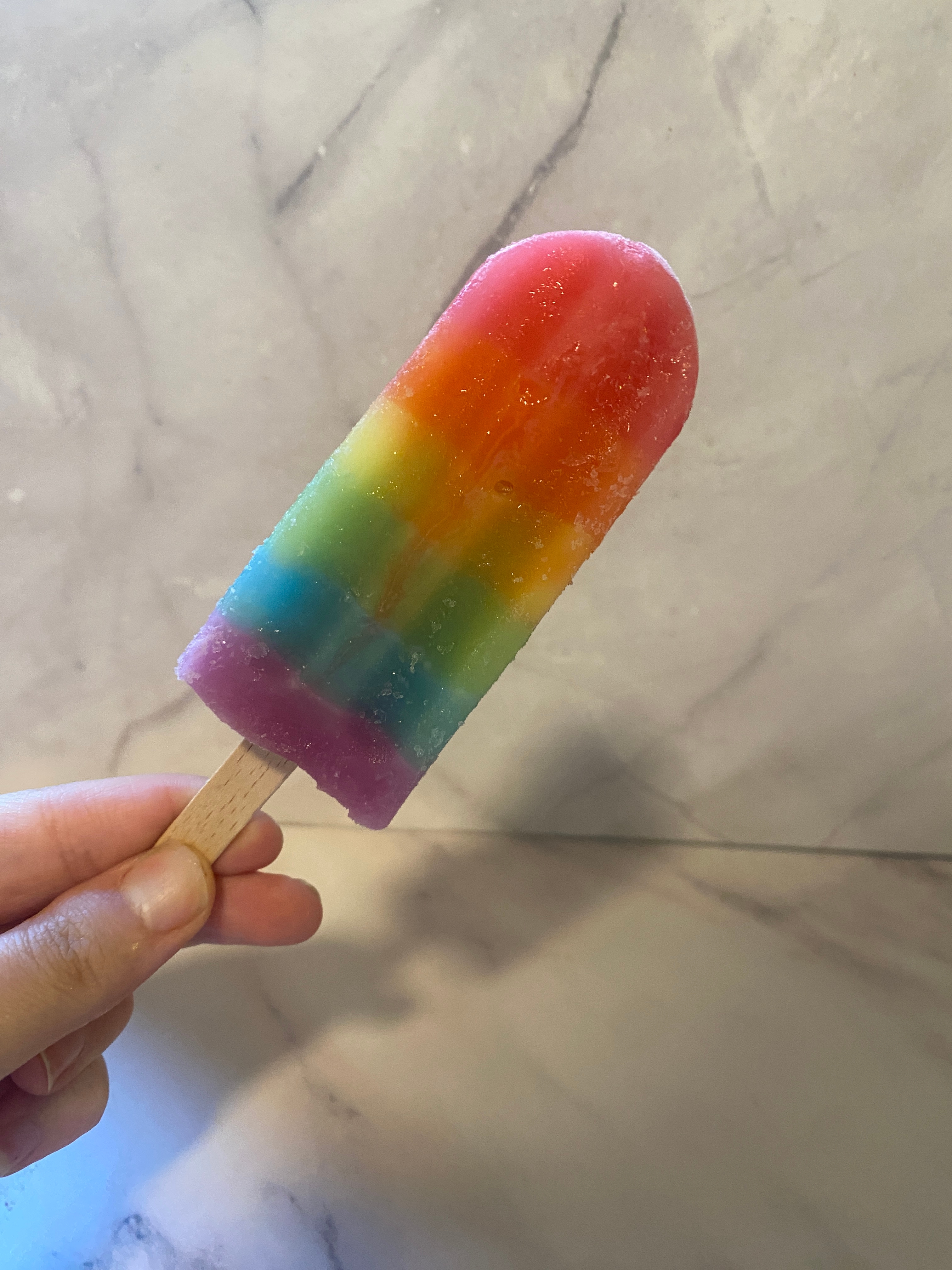 A hand holds a rainbow-colored popsicle against a marble background