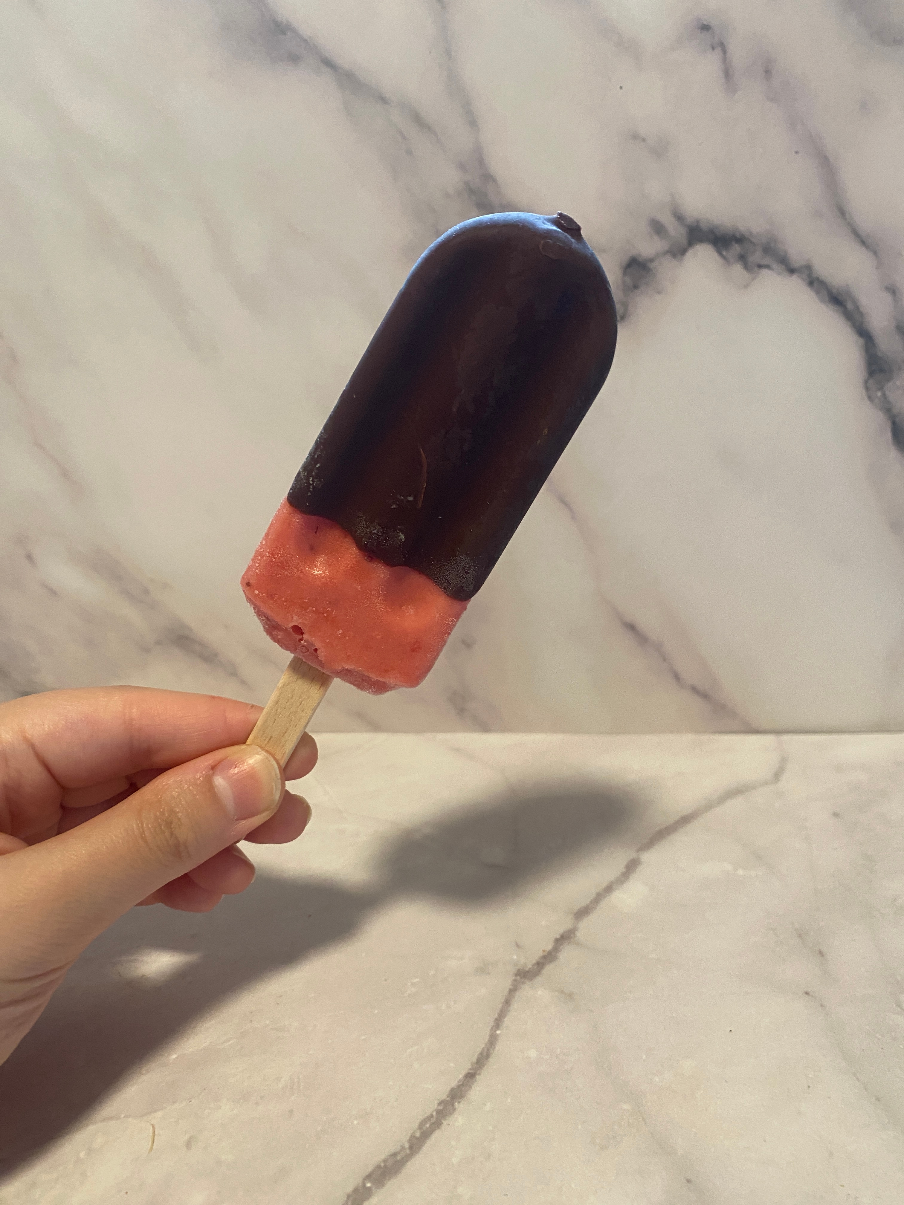 Person holding a chocolate-dipped ice cream bar with a berry-colored base against a marble backdrop