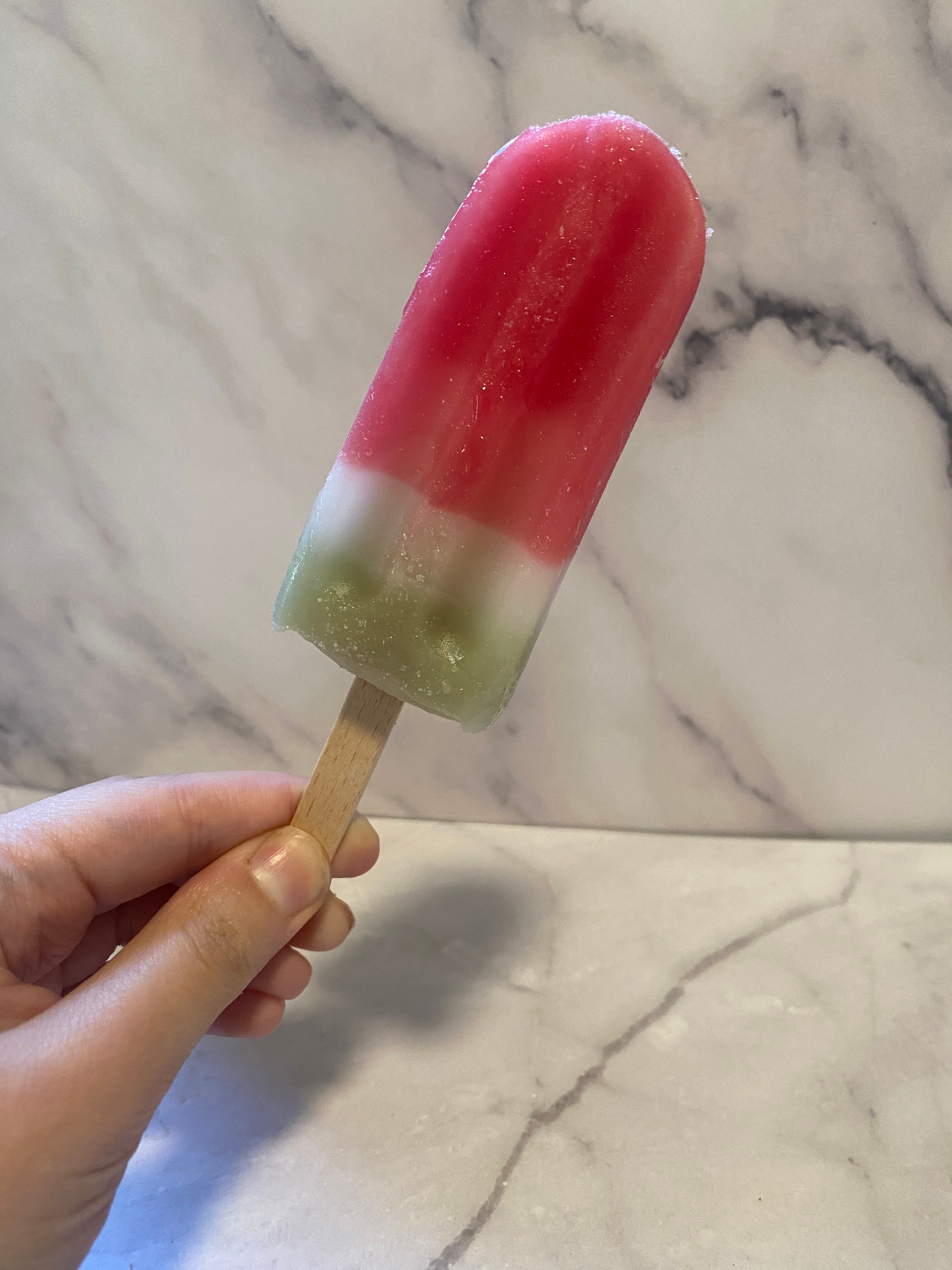 Hand holding a popsicle with three distinct layers against a marble-like background