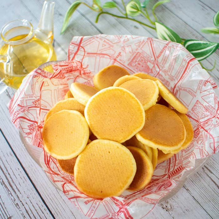 小さなパンケーキがバスケットに盛られ、背景にティーポットと緑の植物が見えます。