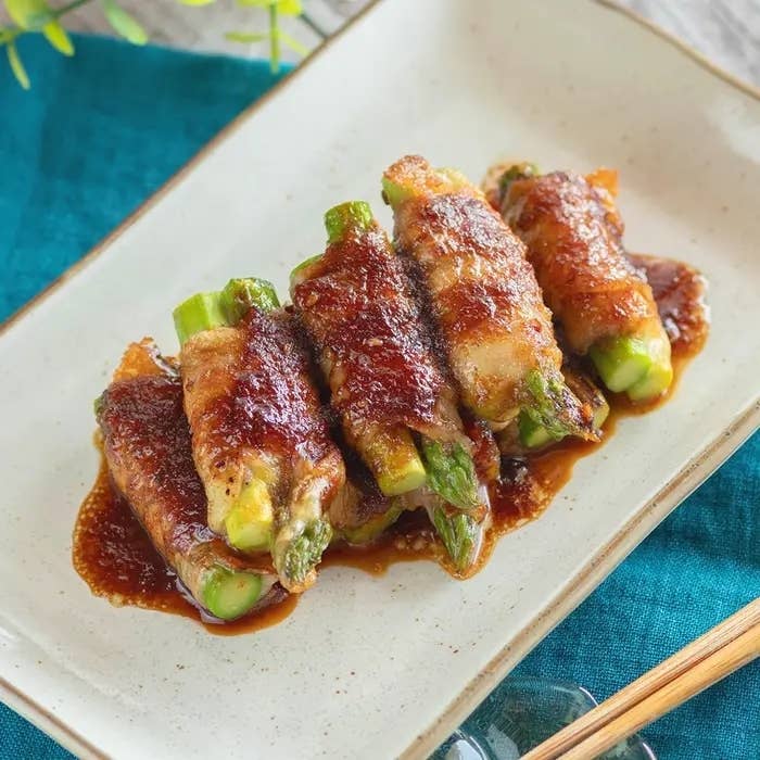 アスパラガスの肉巻きが白い皿に5つ盛られ、ソースがかかっている料理。テーブル上に配置。