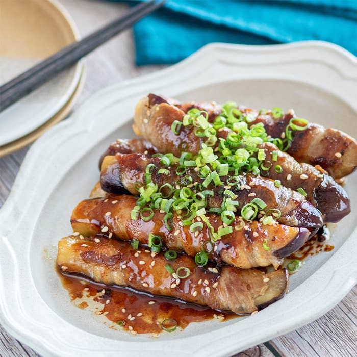 なすと豚肉の巻き料理、細かく刻んだネギとゴマがのった和風ソースがかかっている。白い皿に盛られている。