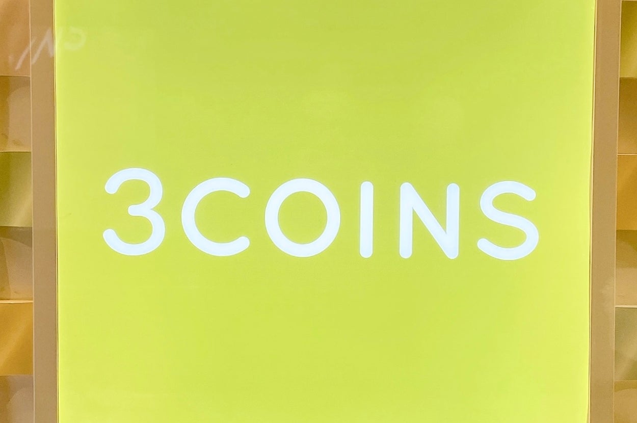「3COINS」のロゴがライトアップされた看板。背景は波打つ模様のあるデザイン。