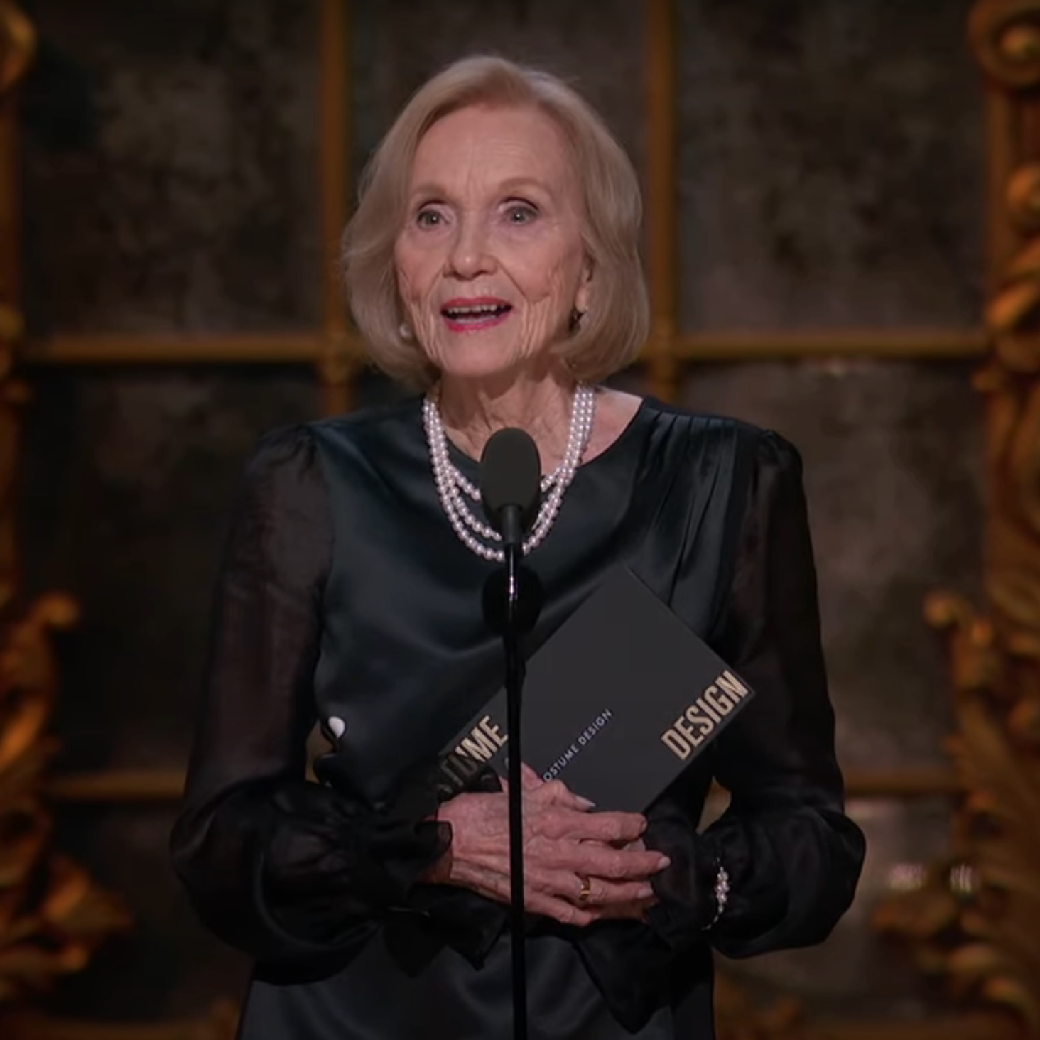 Eva Marie Saint stands at a microphone holding an Oscars envelope, dressed in an elegant black outfit and pearls