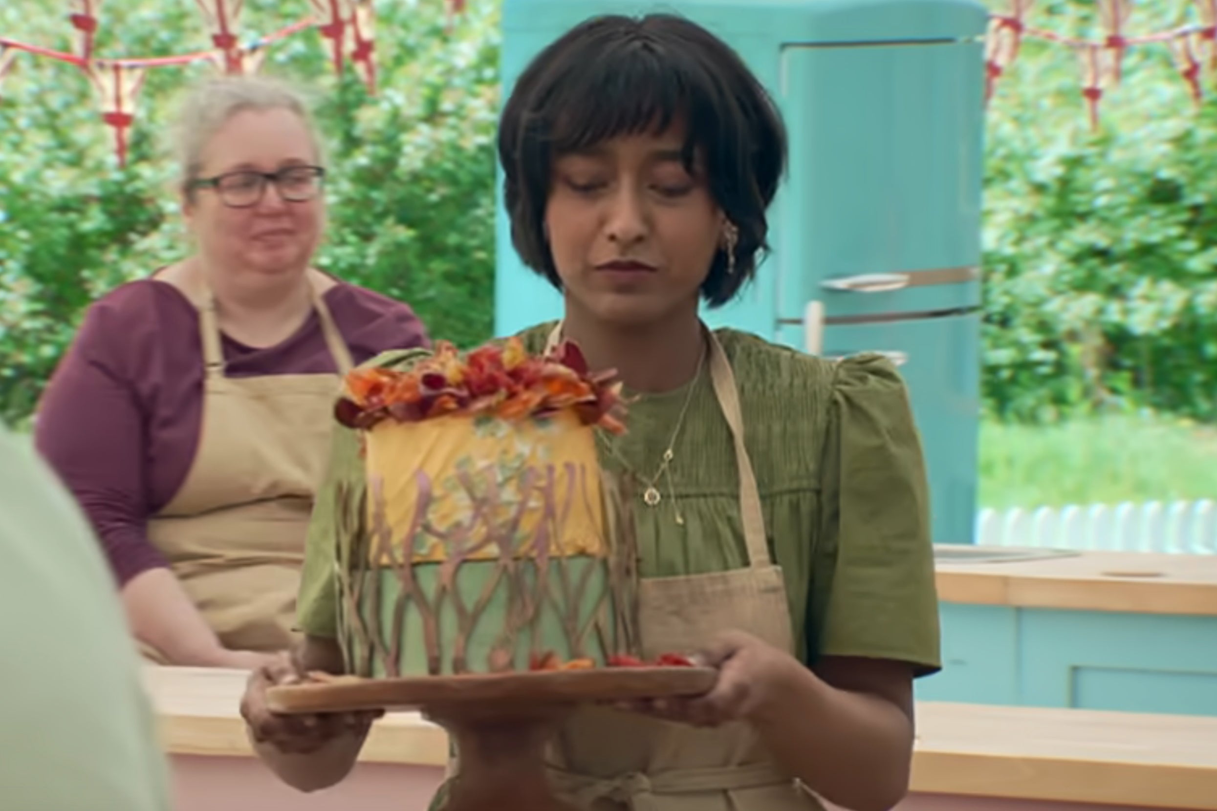 Sumayah from The Great British Baking Show holding a decorated cake with autumn leaves