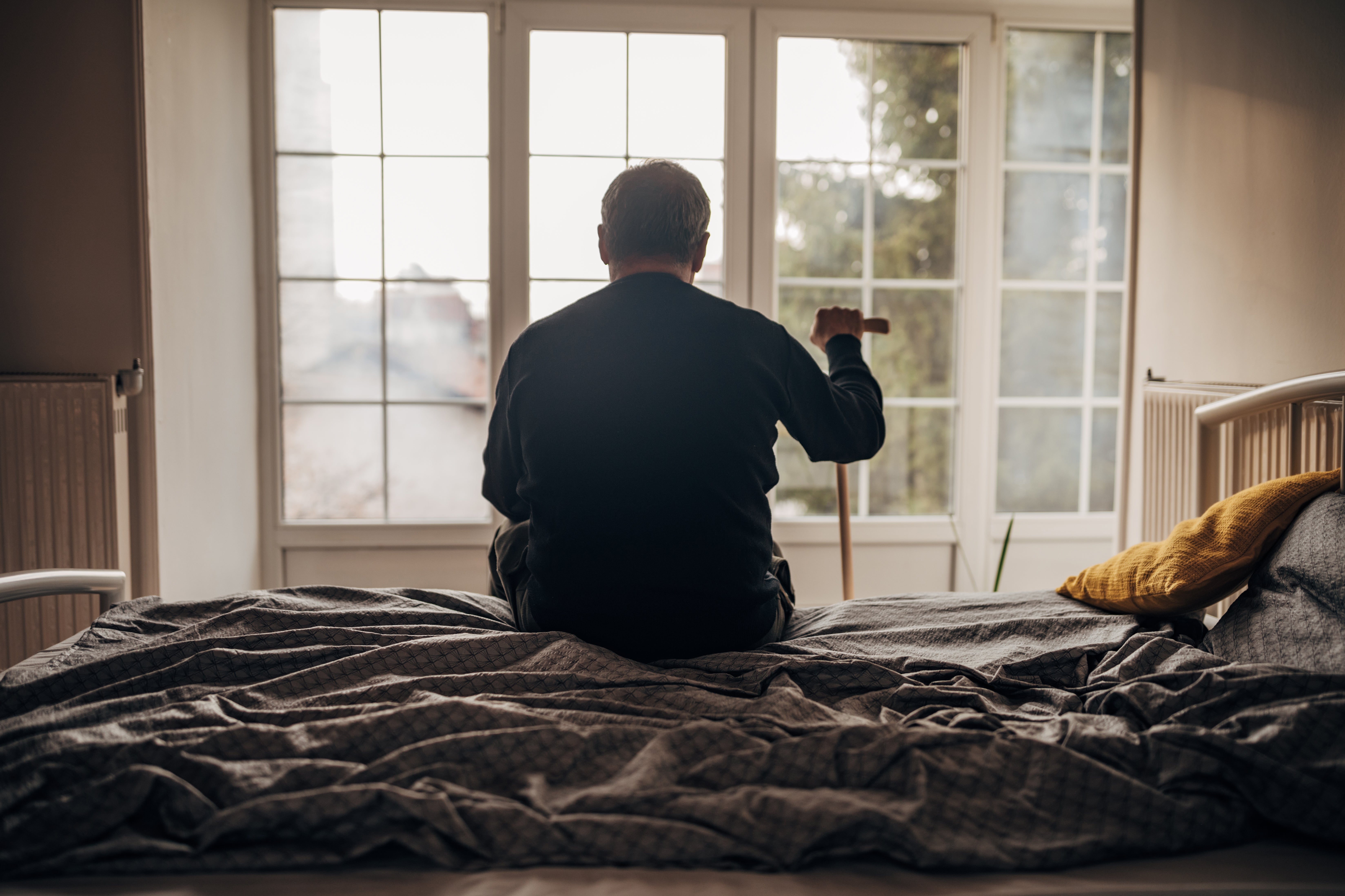 Personen som sitter på sengen med stokk, vendt mot stort vindu. Rommet er mykt opplyst, og skaper en rolig og introspektiv atmosfære