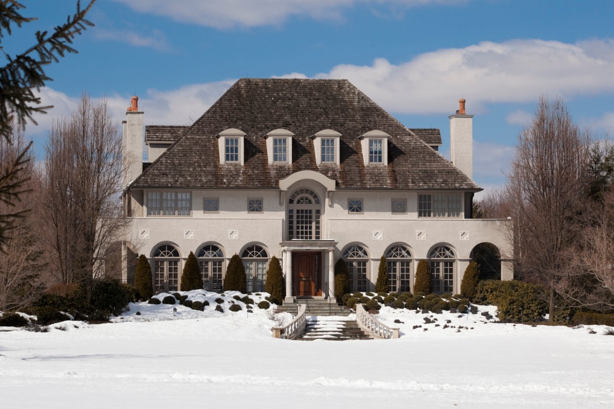 Large, elegant house with steep roof, multiple windows, and arched facade. Snow covers the ground in front, surrounded by leafless trees