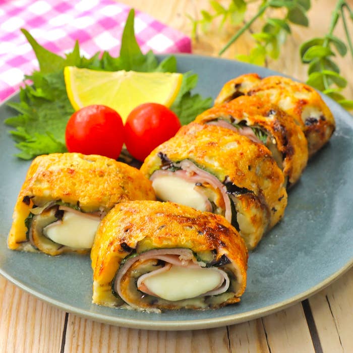 お皿に盛られたチーズとハムを巻いた焼きスフレ風の料理。レモンとトマトが付け合わせされている。