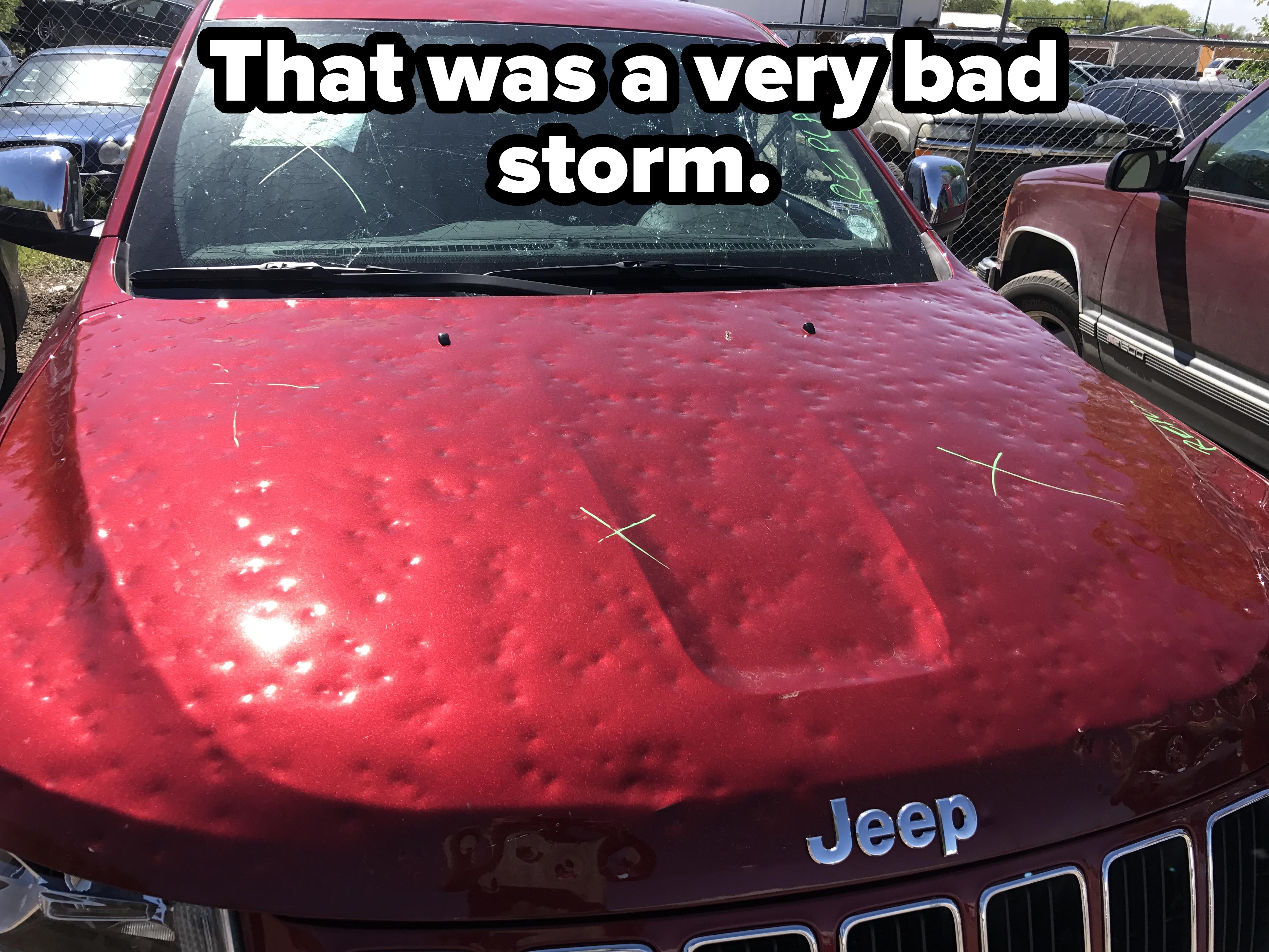 Jeep with a heavily dented hood, likely from hail damage, parked in a lot with other vehicles