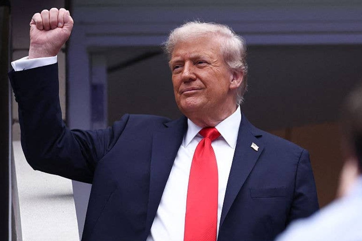 Person in a dark suit and red tie raises a fist in the air, appearing confident and poised at a public event