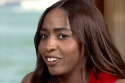 Ayo Edebiri with long hair in a red top speaking in an interview setting, water visible outside the window