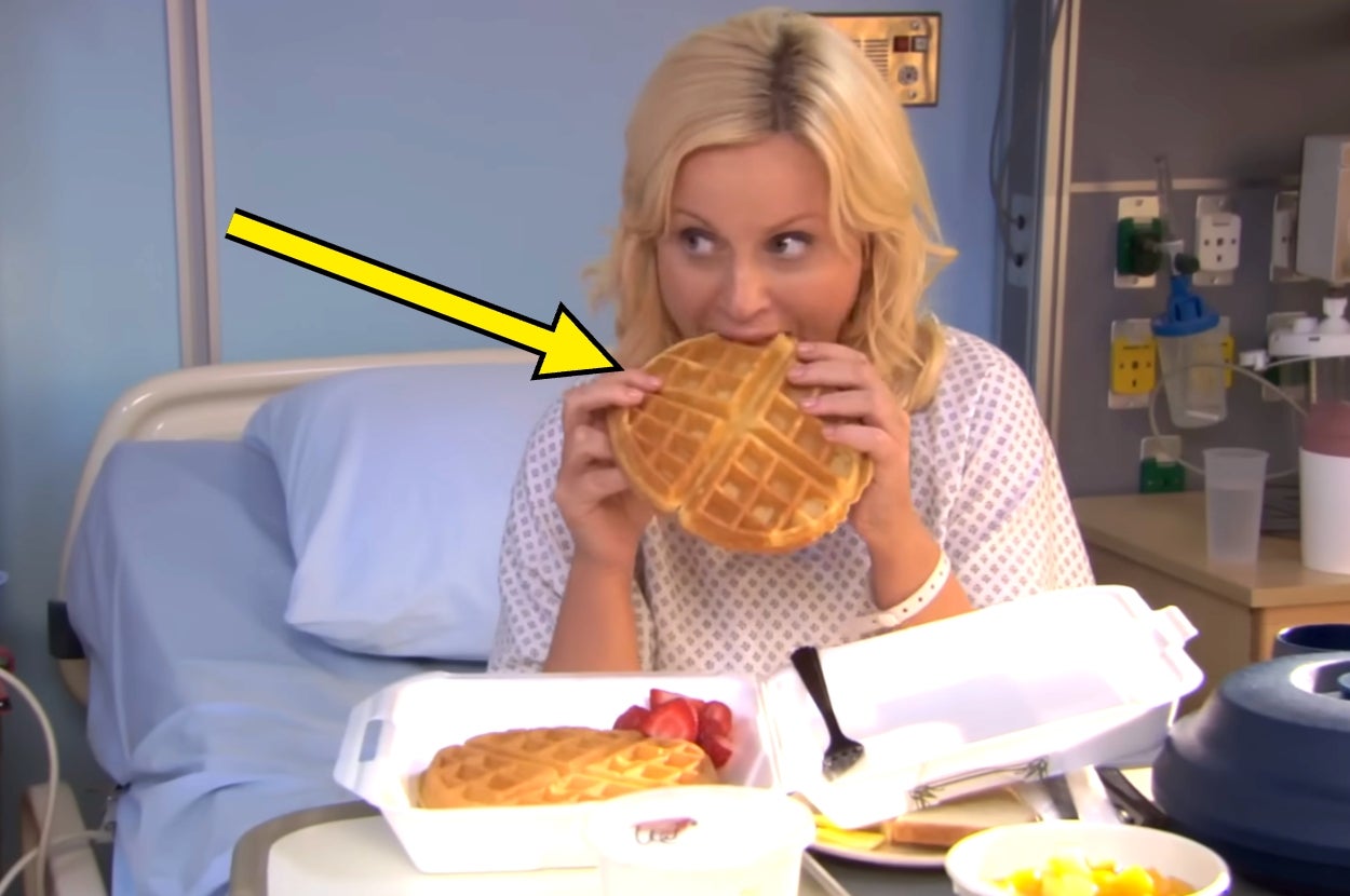 Leslie Knope in a hospital gown enjoys waffles from a takeout box on a bed, with a side of strawberries